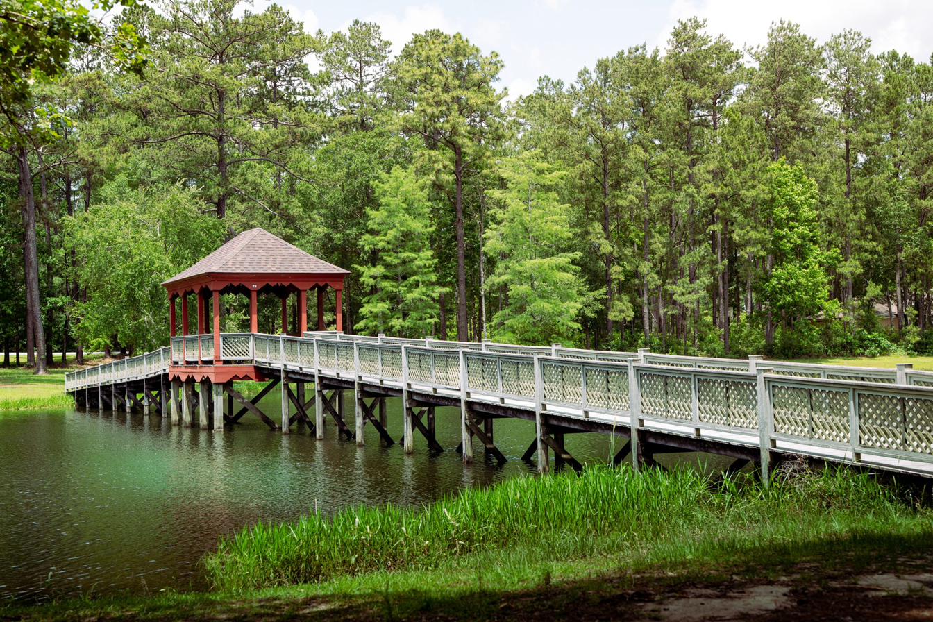 A wooden footbridge crosses the lake at the Solomon Center, with the gazebo visible in golden afternoon light surrounded by Louisiana pine forest